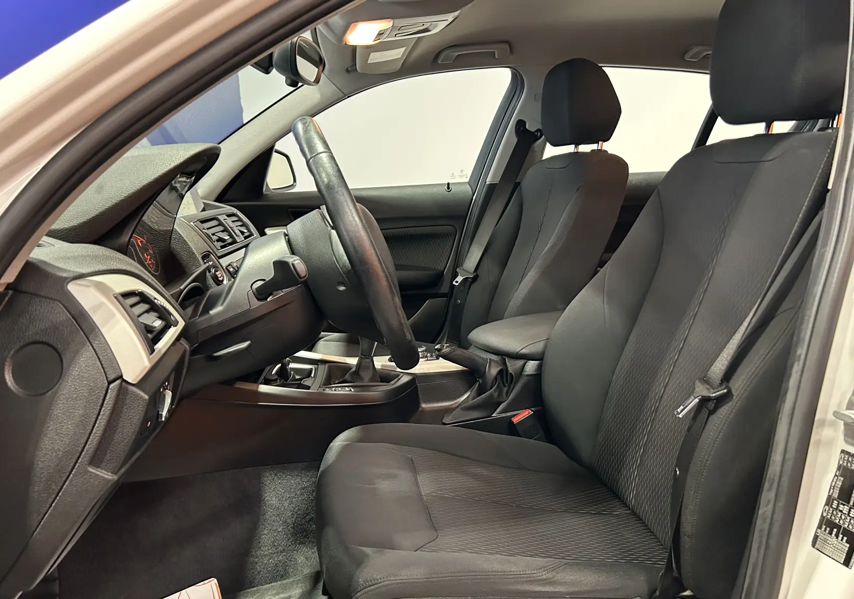 Vue intérieure côté conducteur d'une BMW Série 1 blanche, montrant les sièges noirs et le tableau de bord avec boîte manuelle.