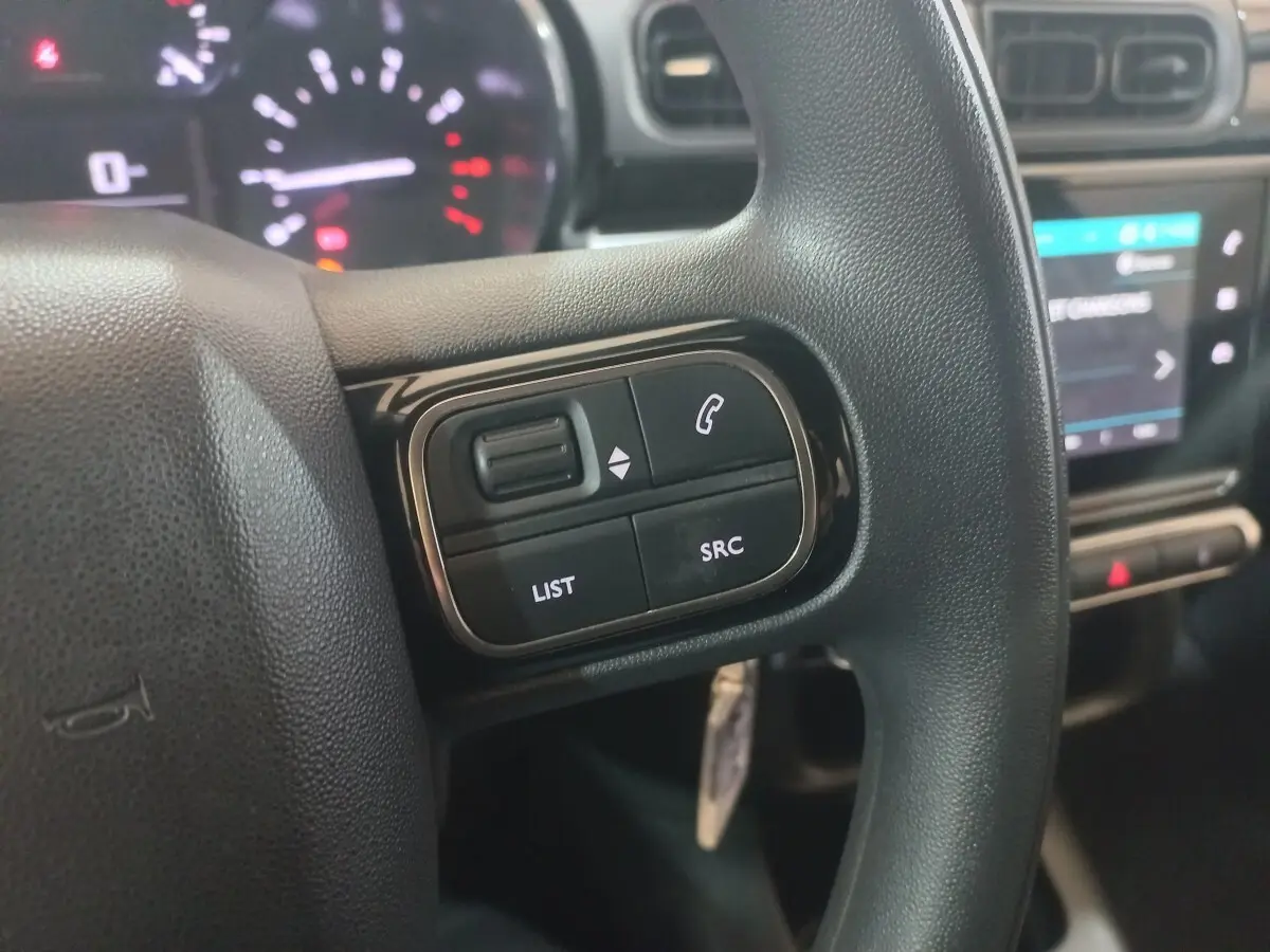 Gros plan sur les commandes gauche du volant noir d'une Citroën C3 2024, avec tableau de bord et écran tactile en arrière-plan.