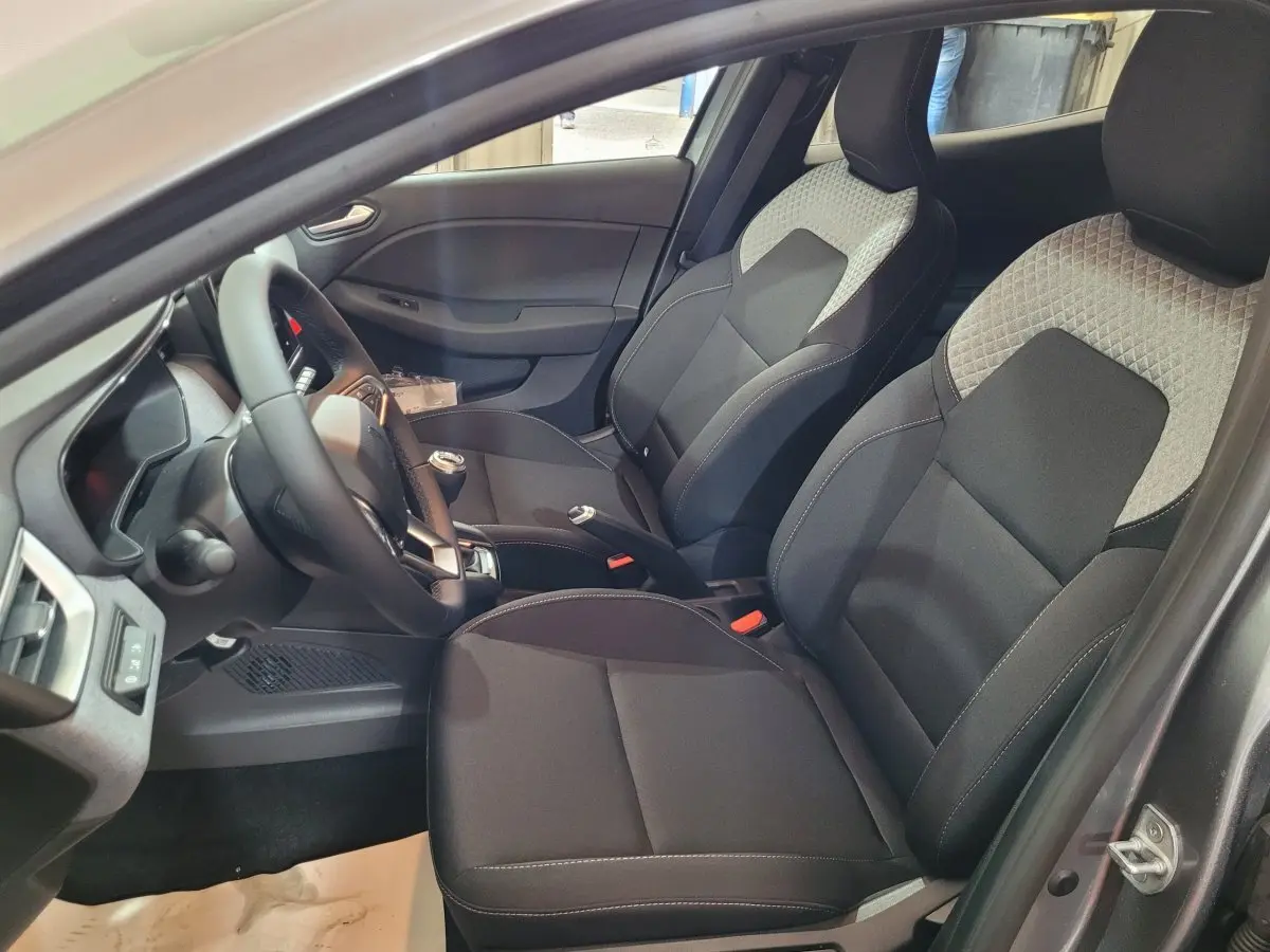 Vue intérieure côté conducteur de la Renault Clio 2025 avec sièges tissu noir et gris, volant multifonctions cuir synthétique.