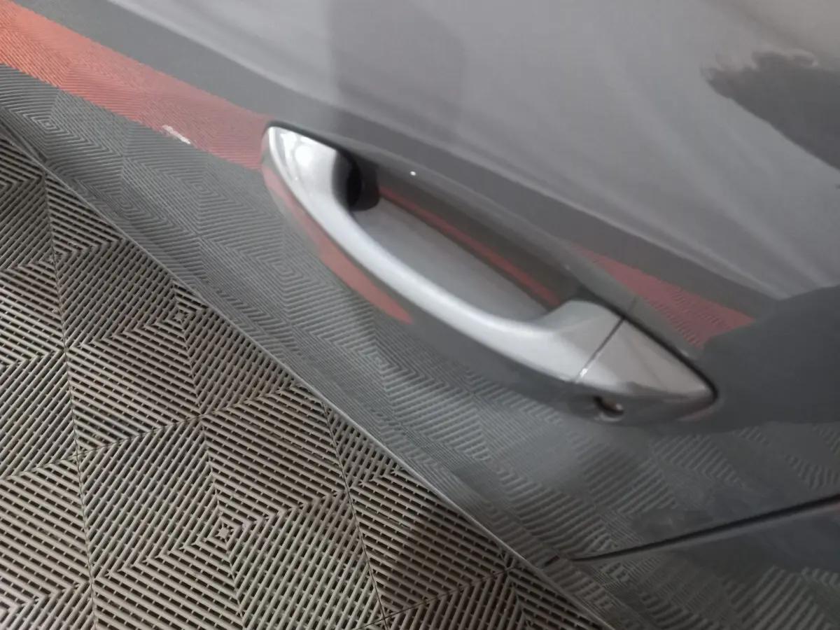 Poignée de porte côté gauche gris foncé de la Peugeot 308 PHEV 180 E-EAT8 Allure 2025, reflet du sol texturé visible.