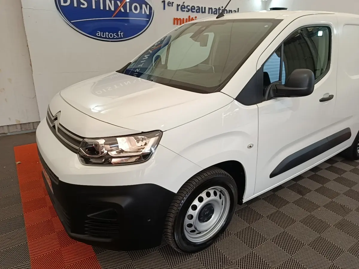 Vue 3/4 avant droite d’un Citroën Berlingo blanc avec pare-chocs noirs et jantes acier en intérieur showroom.