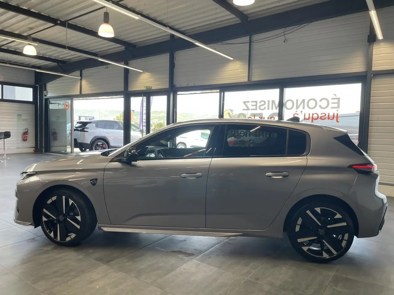 Profil droit de la Peugeot 308 GT 2026 gris Artense avec jantes alliage bi-tons et blason sur aile avant.