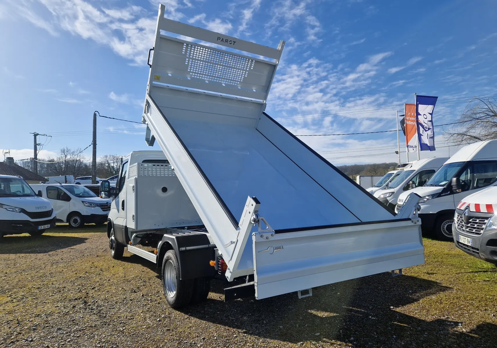 Vue arrière en légère contre-plongée d’un Iveco Daily blanc avec benne acier relevée et coffre latéral visible.