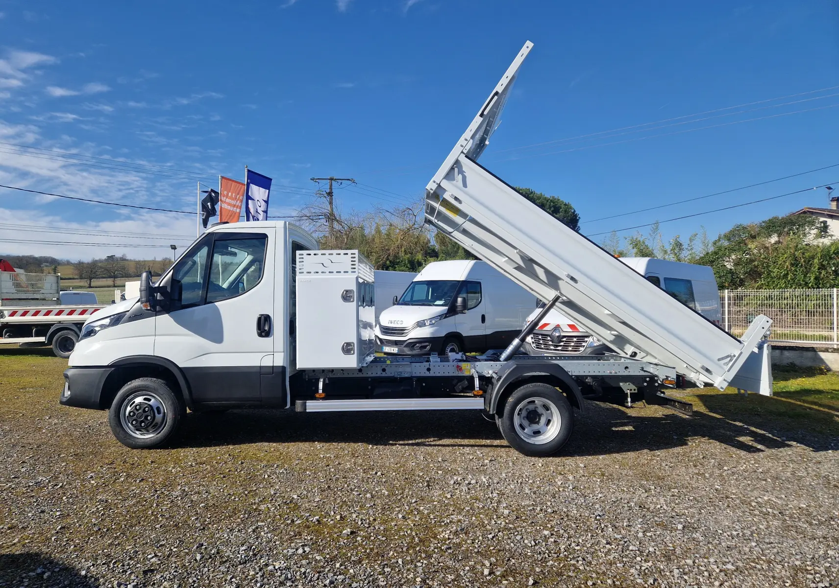 Vue latérale droite d’un Iveco Daily blanc avec benne relevée et coffre latéral acier, sur un terrain extérieur ensoleillé.