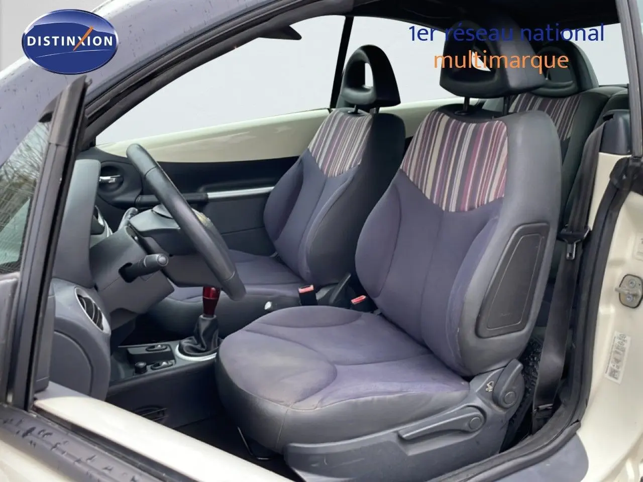 Vue intérieure côté gauche de la Citroën C3 Pluriel ivoire, sièges avant en tissu rayé noir et beige avec volant cuir.