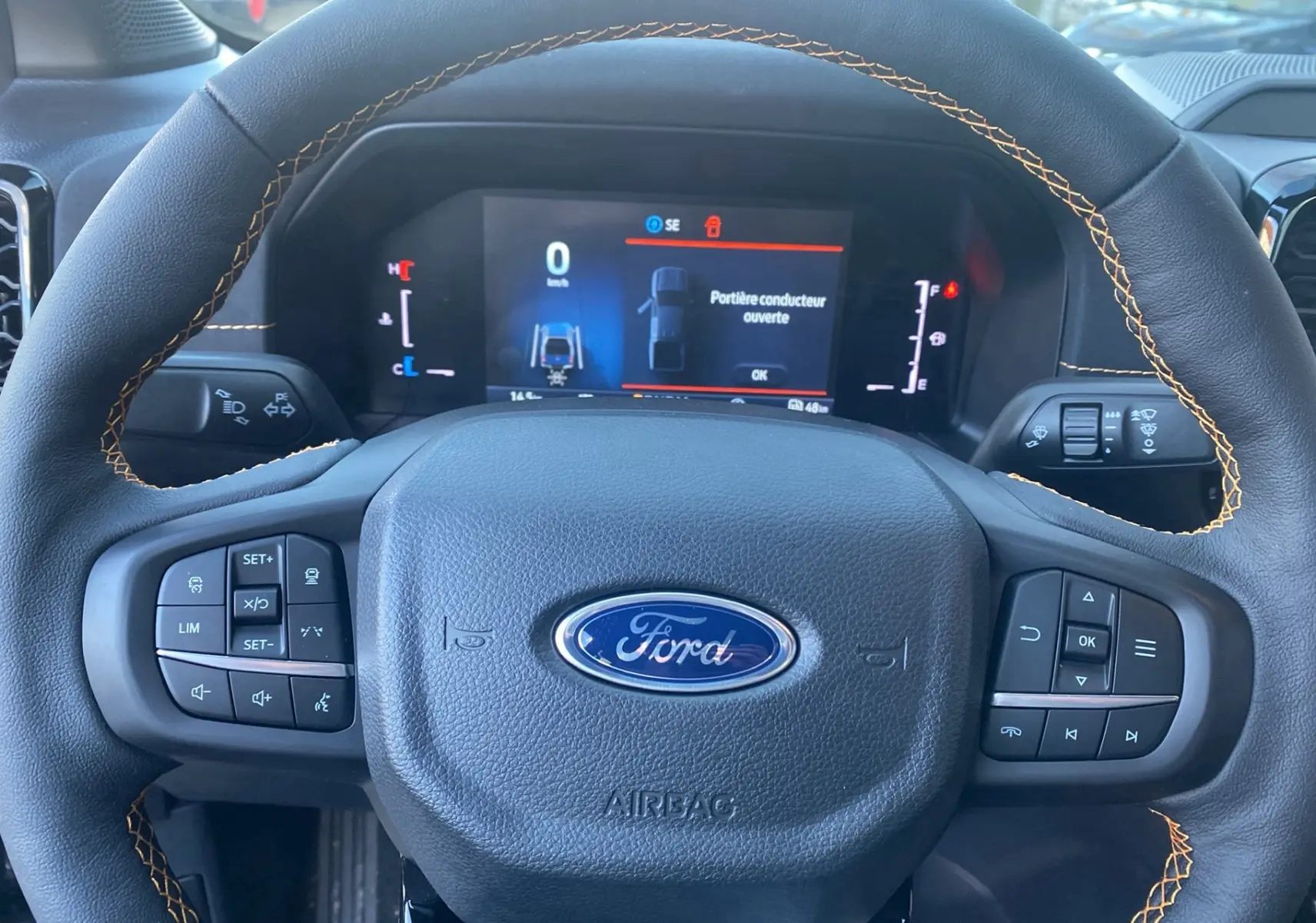 Vue rapprochée du volant en cuir noir avec surpiqûres orange du Ford Ranger 2025, tableau de bord digital allumé.