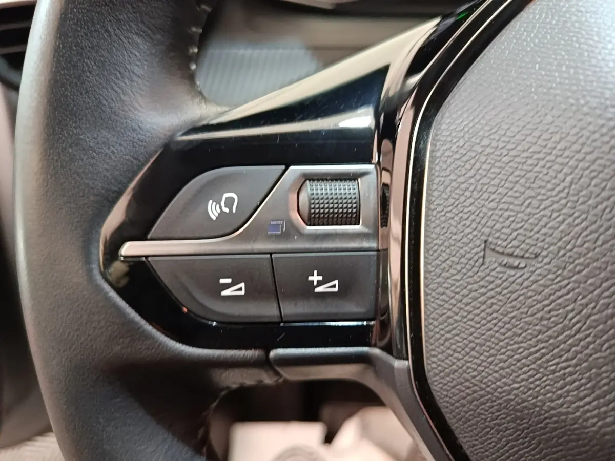 Gros plan sur les commandes gauche du volant cuir noir du Peugeot 208 gris clair, boutons multifonctions et molette.