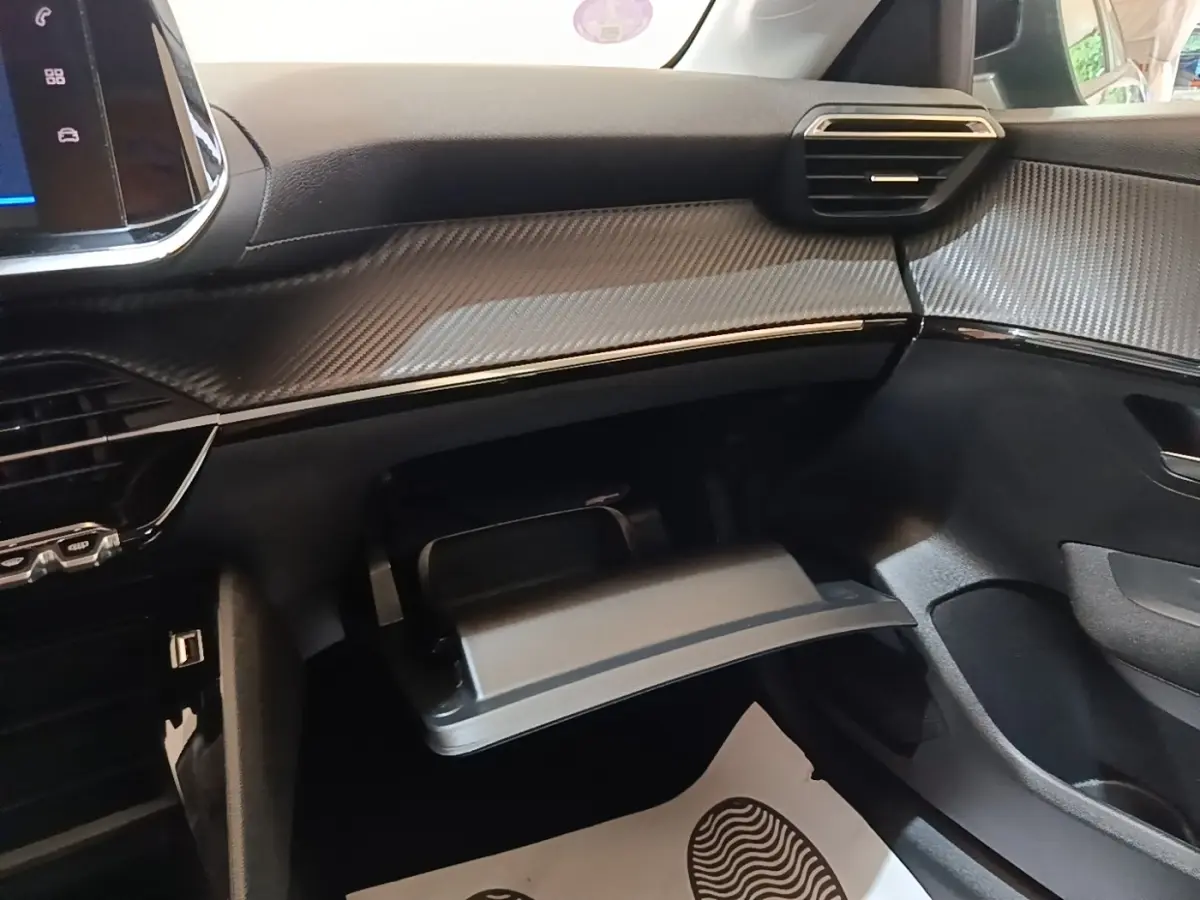 Vue intérieure côté passager du tableau de bord gris foncé avec boîte à gants ouverte et finition effet carbone sur Peugeot 208 2023.