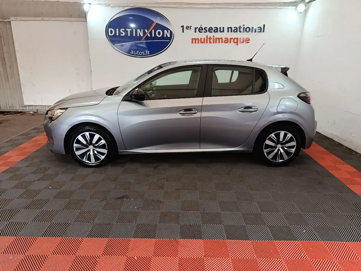 Profil droit d'une Peugeot 208 gris clair 2023 garée en intérieur sur sol à damiers noir et rouge.