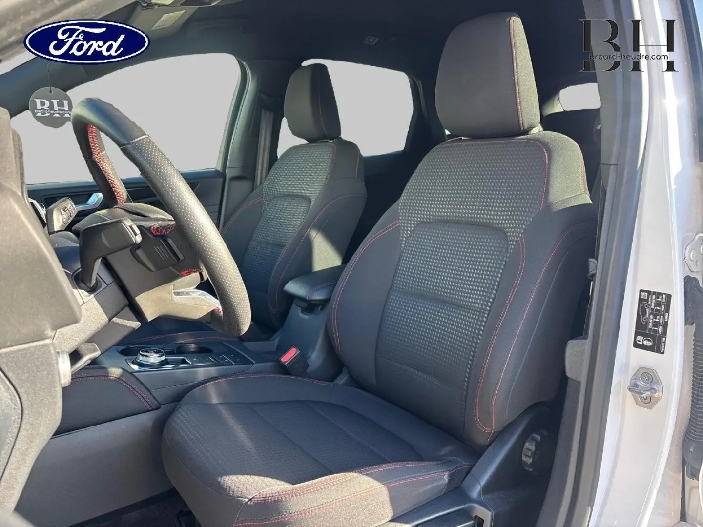 Vue intérieure côté conducteur du Ford Kuga blanc, sièges sport noirs avec surpiqûres rouges et volant cuir multifonction.