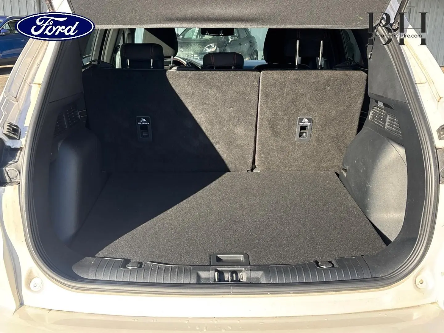 Vue arrière du coffre ouvert d'un Ford Kuga blanc Premium Platinum, montrant la banquette arrière et le tapis de coffre noir.