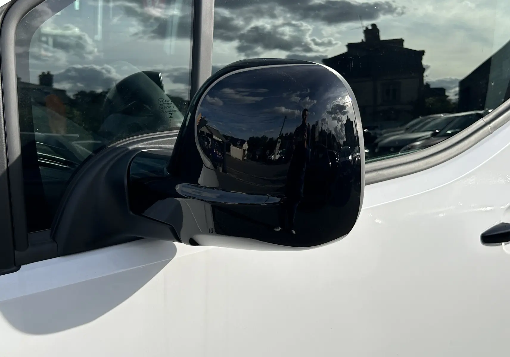 Rétroviseur noir en gros plan côté gauche d'un Peugeot Partner Fourgon blanc, reflet d'un ciel nuageux et d'un bâtiment.