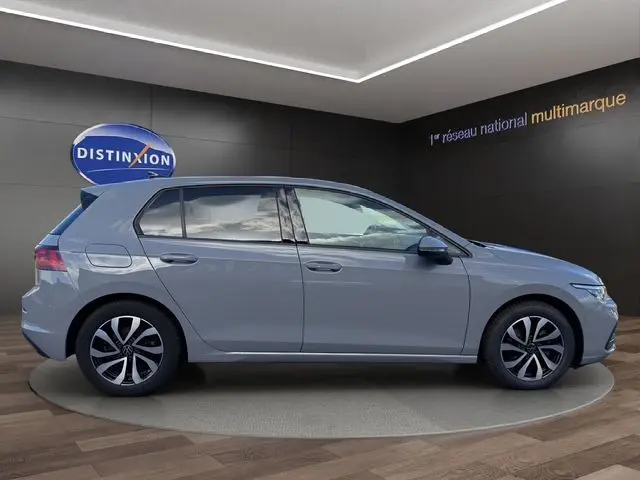 Vue de profil côté gauche d'une Volkswagen Golf VIII gris lunaire avec jantes alu et toit panoramique visible en intérieur showroom.