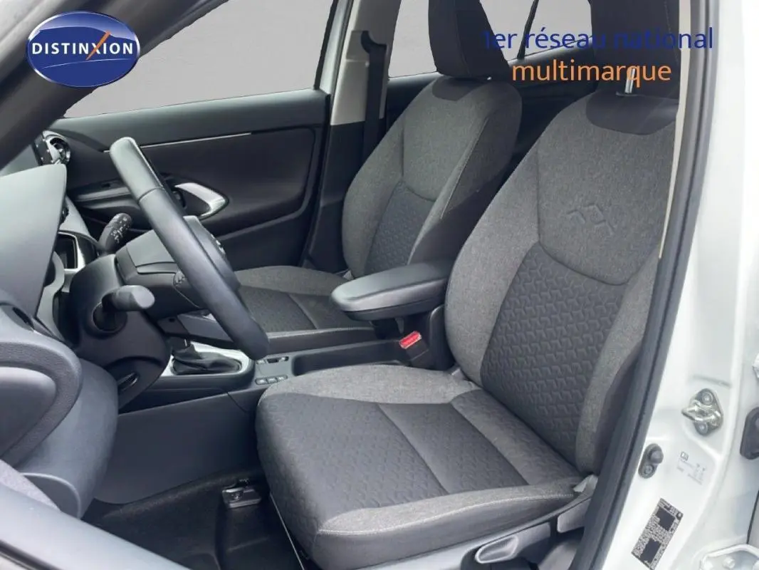 Vue intérieure côté conducteur de la Toyota Yaris Cross Hybrid blanche, sièges tissu gris et volant cuir visible.