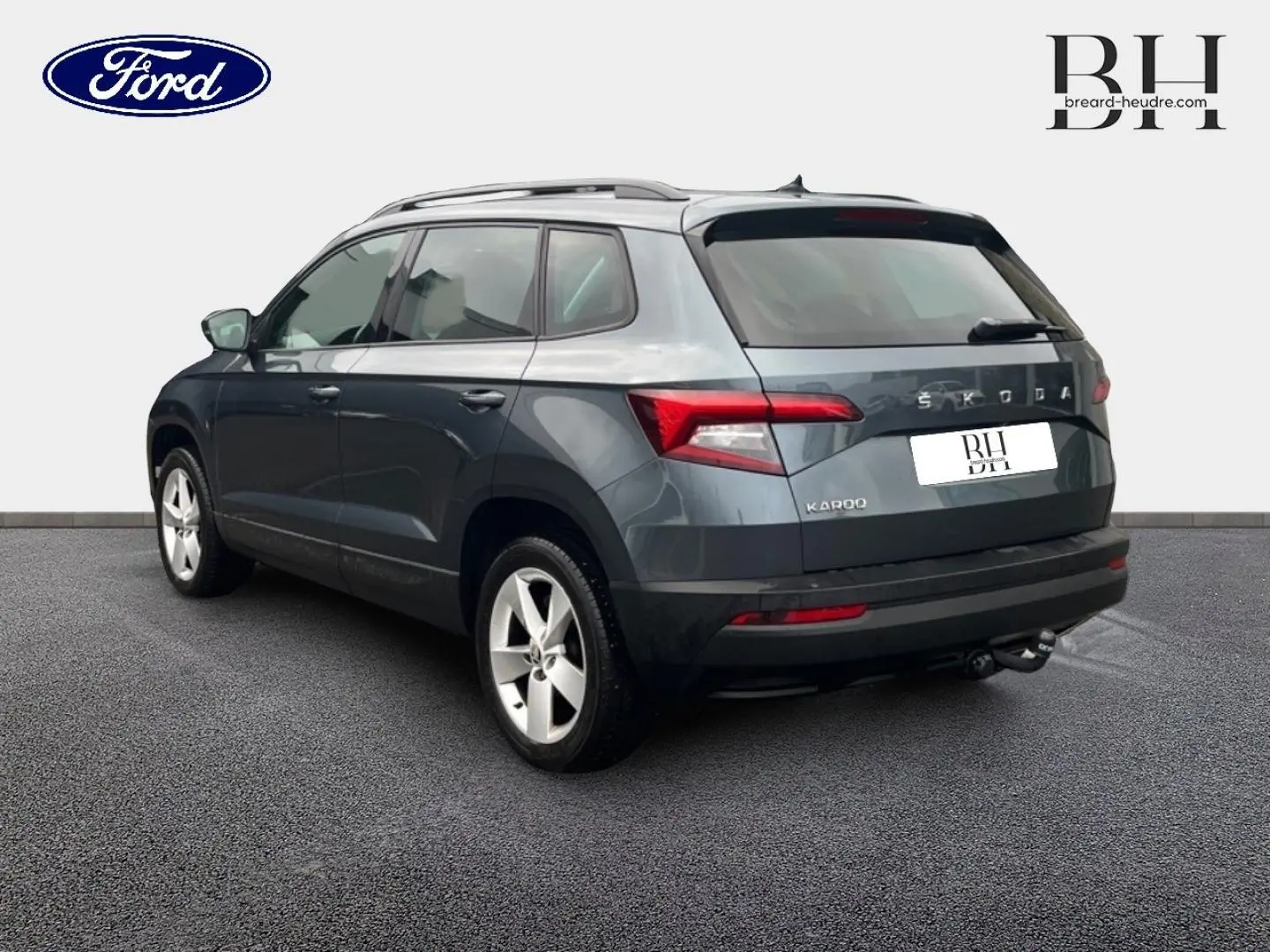 Vue 3/4 arrière droite d'un Skoda Karoq gris Business avec jantes alu et attelage visible sur fond neutre.
