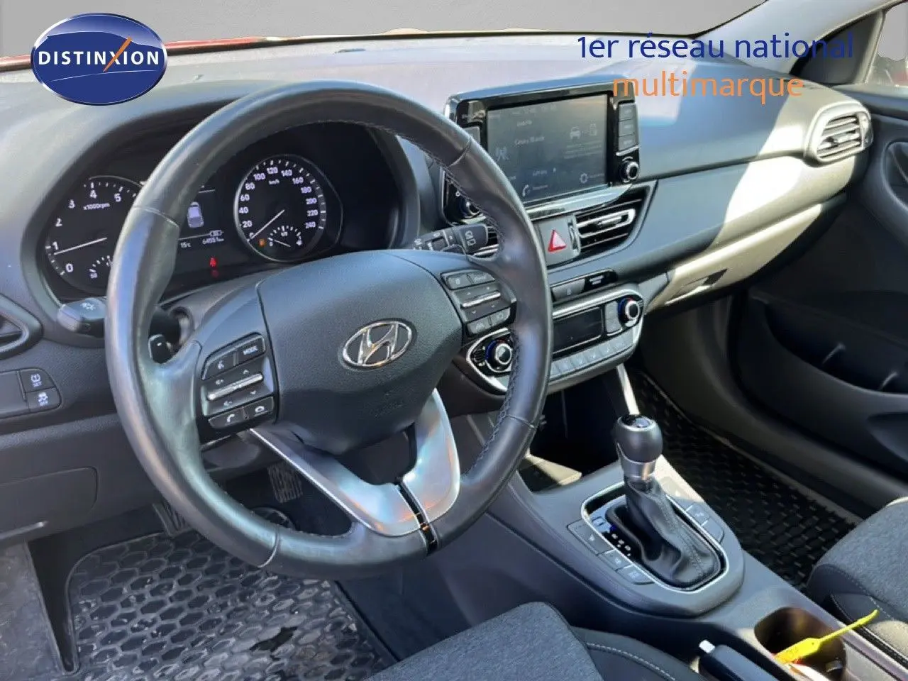 Vue intérieure du poste de conduite de la Hyundai i30 SW 2023, volant cuir noir et console centrale avec boîte automatique.