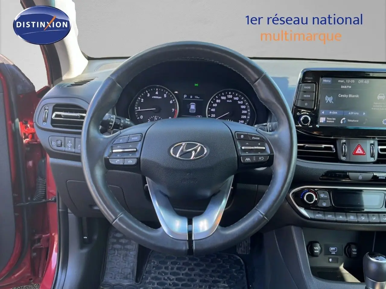 Vue intérieure centrée sur le volant et le tableau de bord d'une Hyundai i30 SW rouge avec écran tactile et commandes au volant.