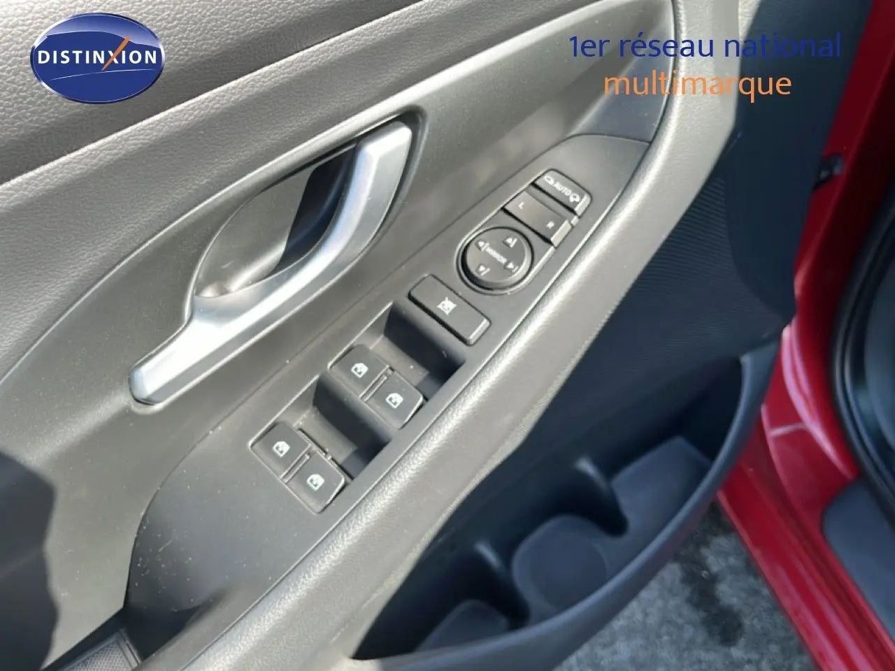Gros plan sur la porte avant gauche d'une Hyundai i30 SW rouge, montrant les commandes des vitres et la poignée intérieure argentée.