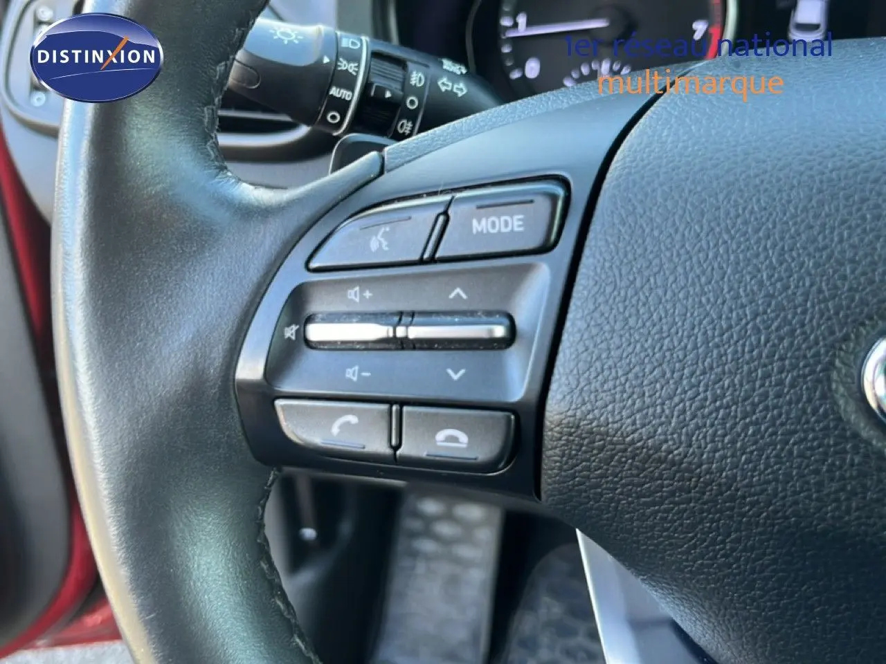 Gros plan sur le volant noir de la Hyundai i30 SW 2023, avec commandes audio et intérieur en rouge Sunset Metal visible en arrière-plan.
