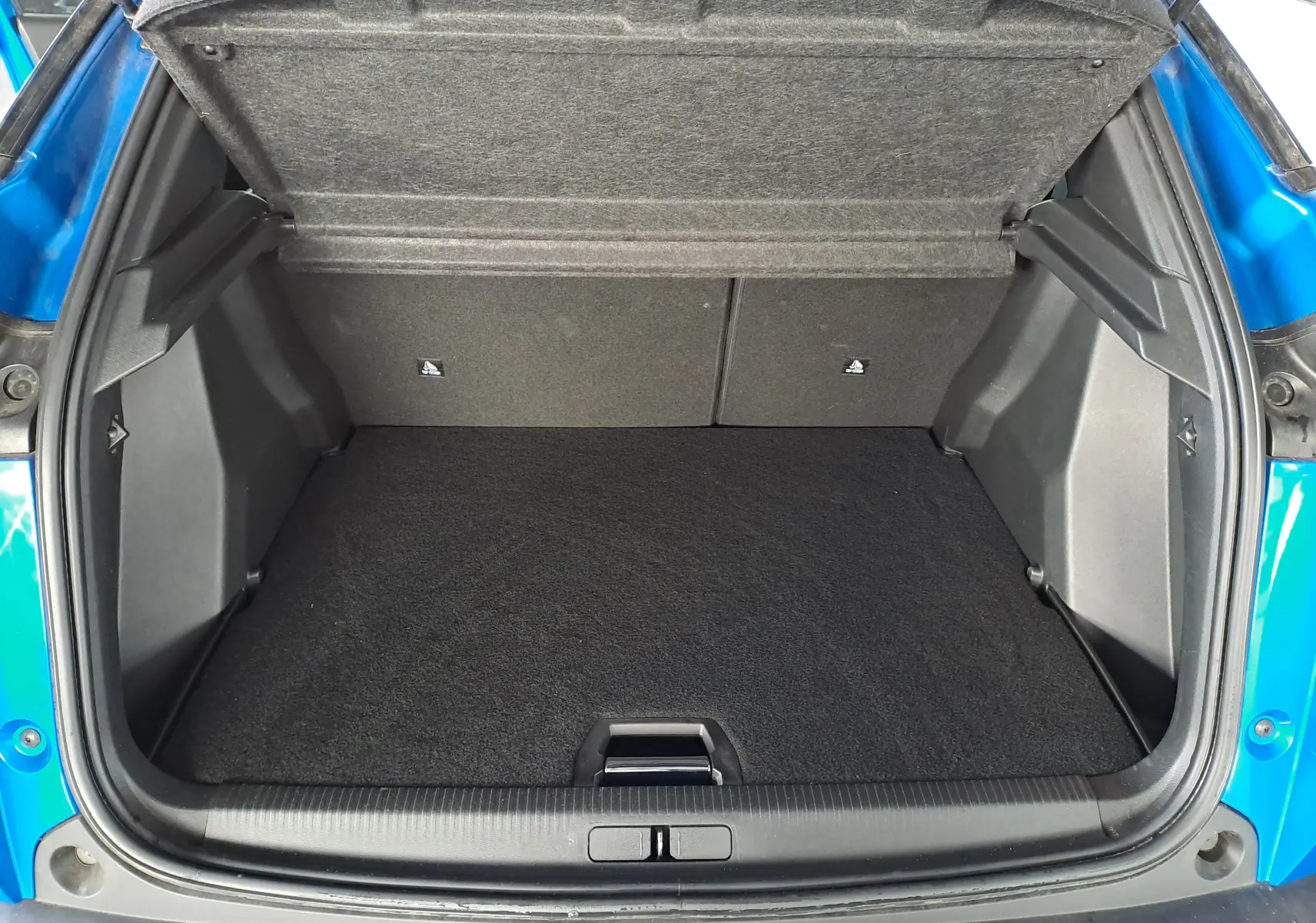 Coffre ouvert vu de l'arrière d'une Peugeot 2008 bleu Vertigo, montrant l'espace de chargement vide et la moquette noire.