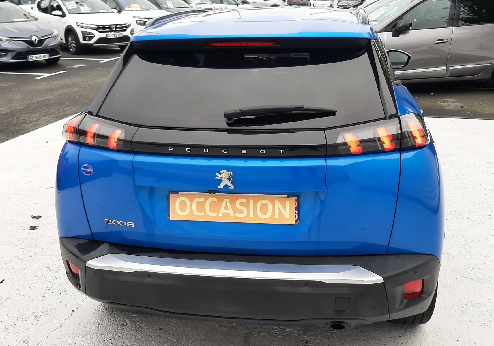 Vue arrière d'un Peugeot 2008 bleu Vertigo avec feux arrière à griffes et plaque "OCCASION" visible sur un parking.