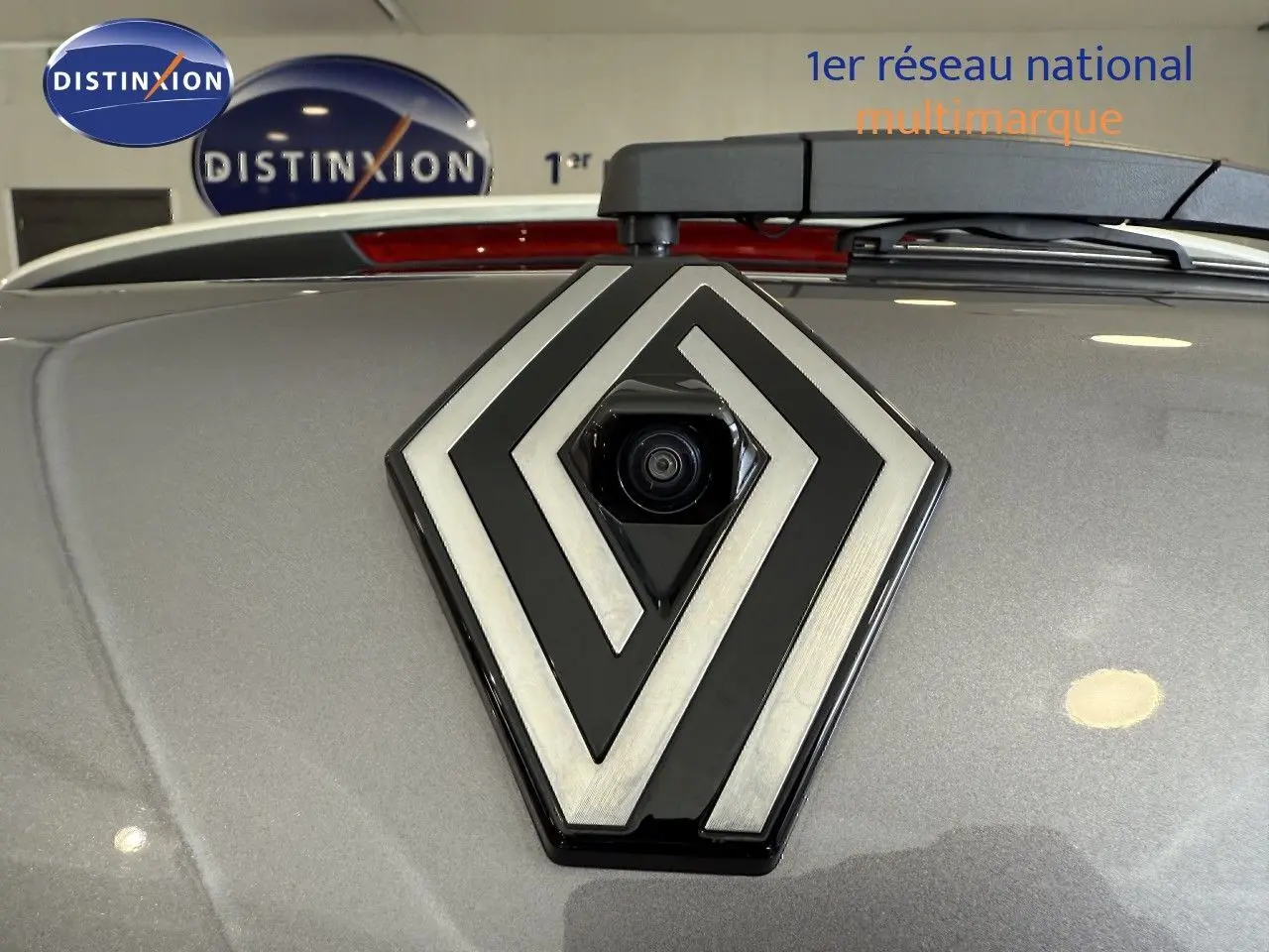 Gros plan sur le logo Renault gris avec caméra intégrée, sur le capot gris cassiopée métallisé du Captur 2025.