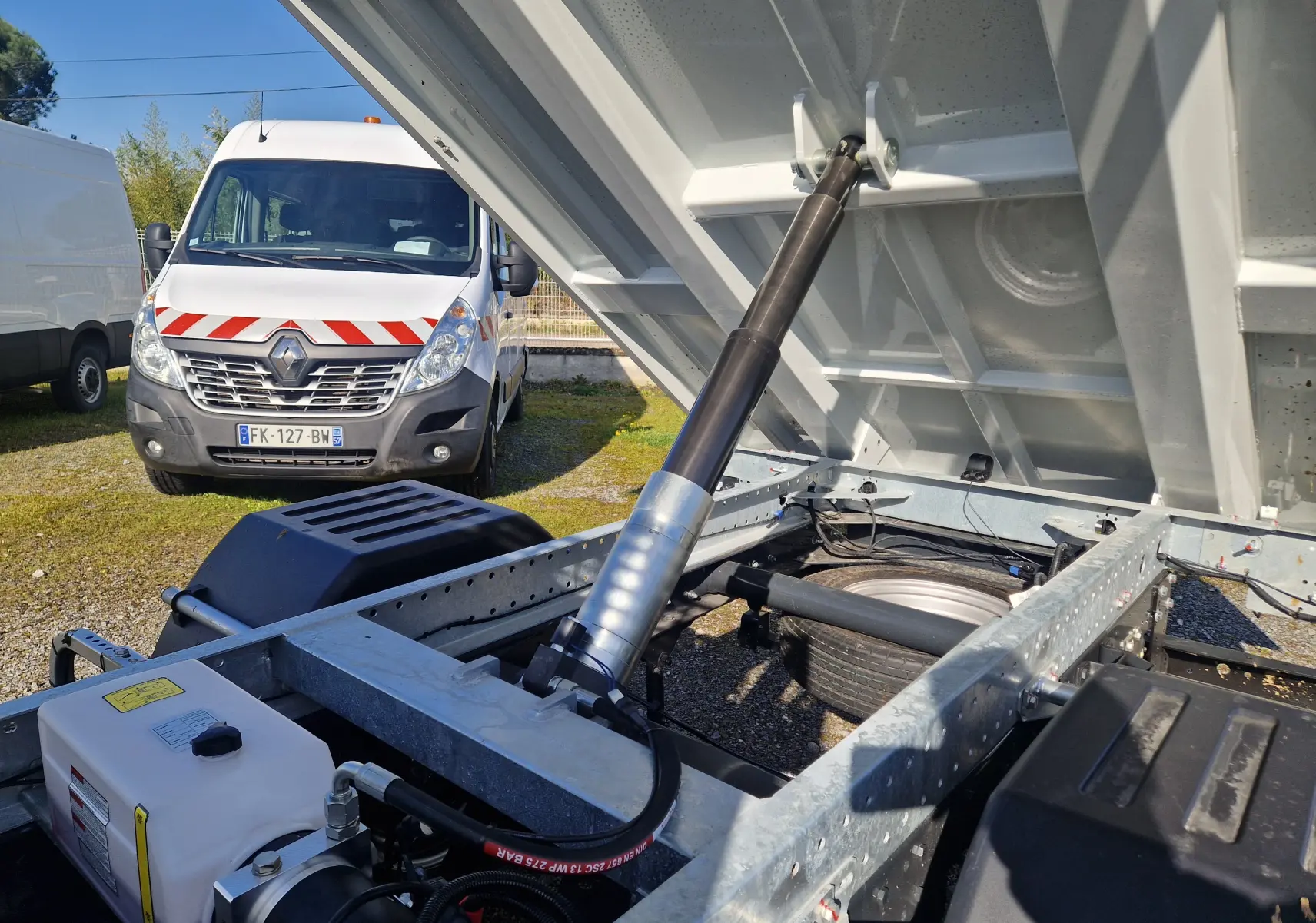 Vue rapprochée du châssis et du système hydraulique de la benne blanche d’un Iveco Daily, avec un fourgon blanc en arrière-plan.