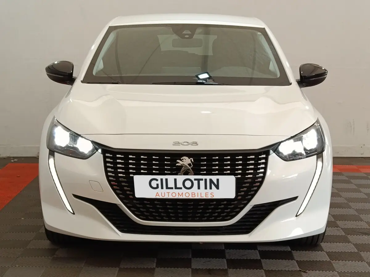 Vue avant d'une Peugeot 208 blanche 2023 avec phares LED allumés et calandre noire distinctive.