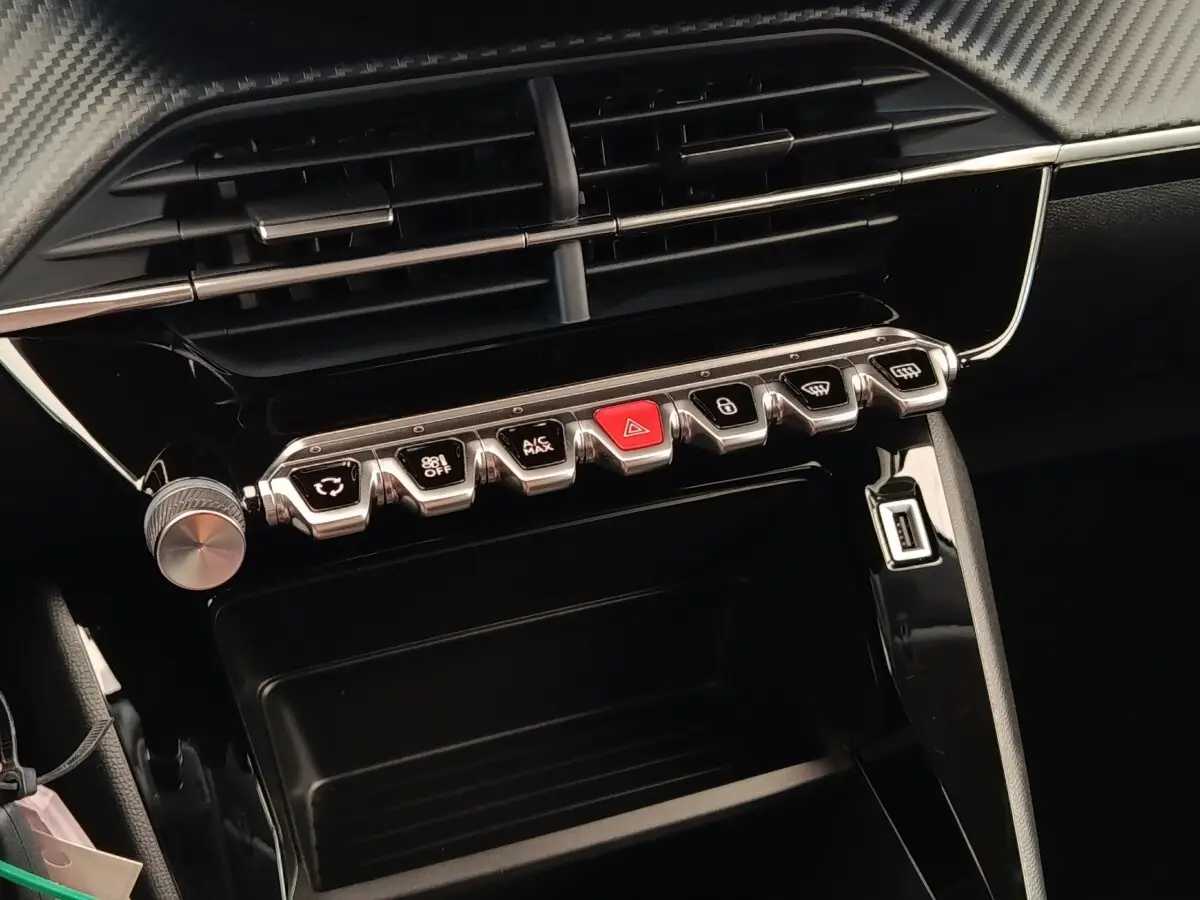 Vue rapprochée du tableau de bord central noir brillant de la Peugeot 208 2023 avec rangée de boutons chromés et bouton rouge d’alerte.