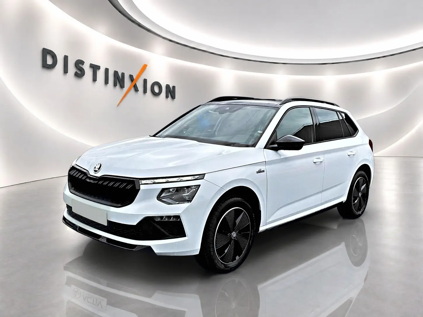 Vue 3/4 avant d'un Skoda Kamiq blanc nacré avec jantes noires et toit panoramique en showroom lumineux.