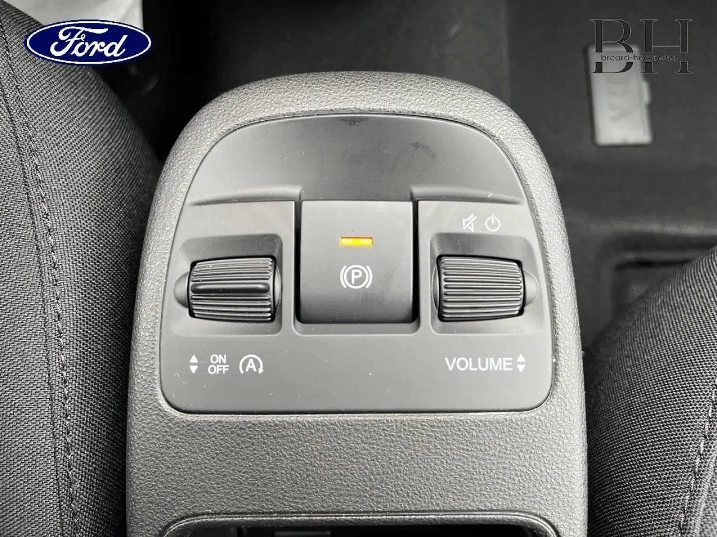 Image montrant le panneau de commandes centrales avec boutons de volume et frein à main électrique, intérieur tissu noir.