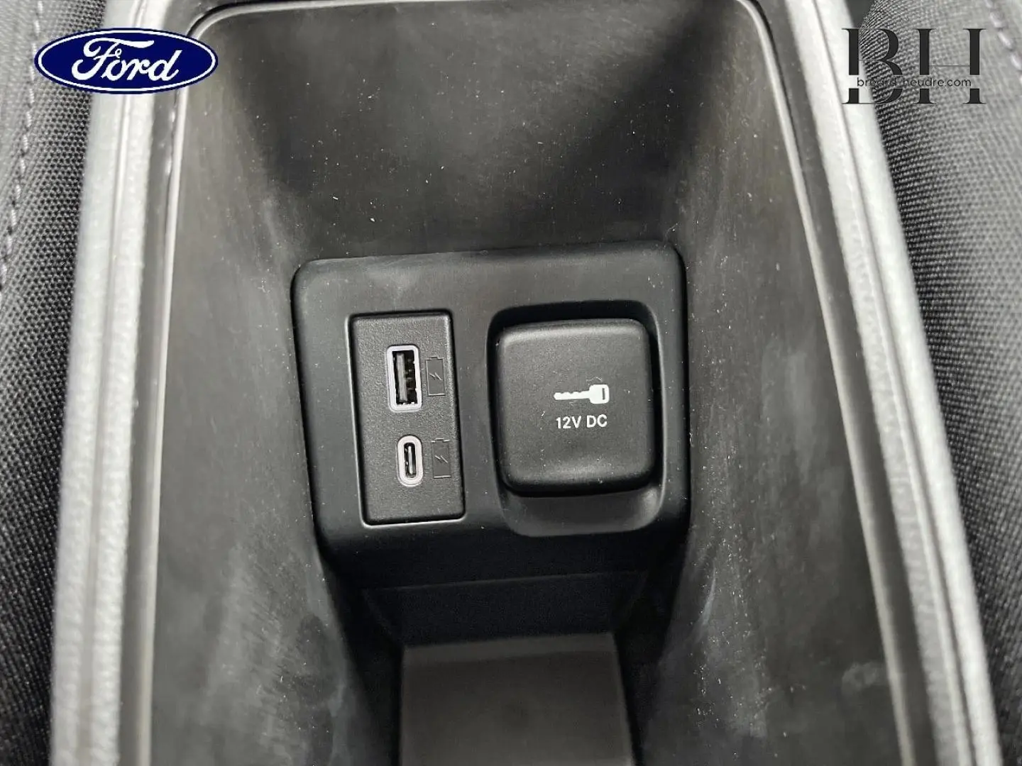 Gros plan sur la prise 12V et ports USB dans le rangement central d’un véhicule, intérieur noir.