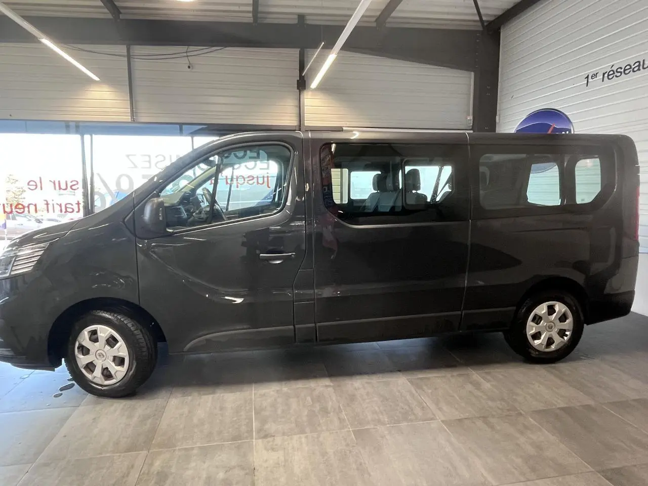 Vue de profil côté gauche du Renault Trafic Fourgon gris foncé avec vitres teintées et jantes 16 pouces en intérieur showroom.