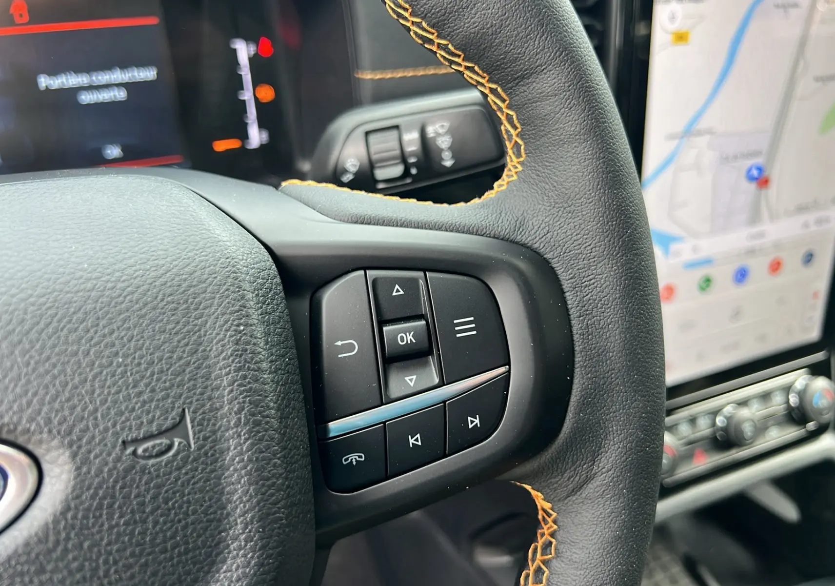 Gros plan sur le volant cuir noir avec surpiqûres orange du Ford Ranger 2026, écran tactile GPS visible en arrière-plan.
