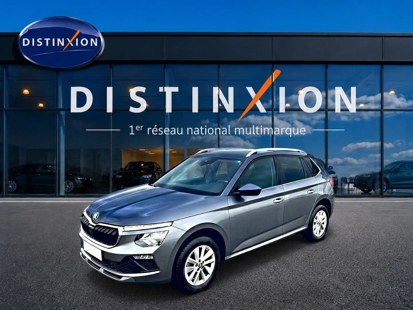 SKODA KAMIQ gris en 3/4 avant droit, avec calandre chromée et barres de toit blanches visibles.