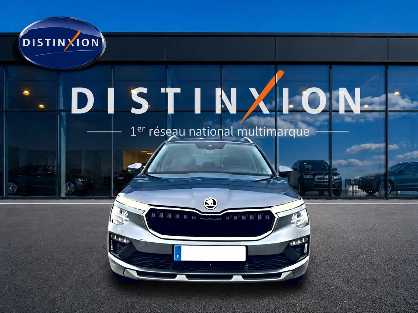 Vue de face d'un Skoda Kamiq gris 2025 avec calandre noire et barres de toit devant une vitrine Distinxion.