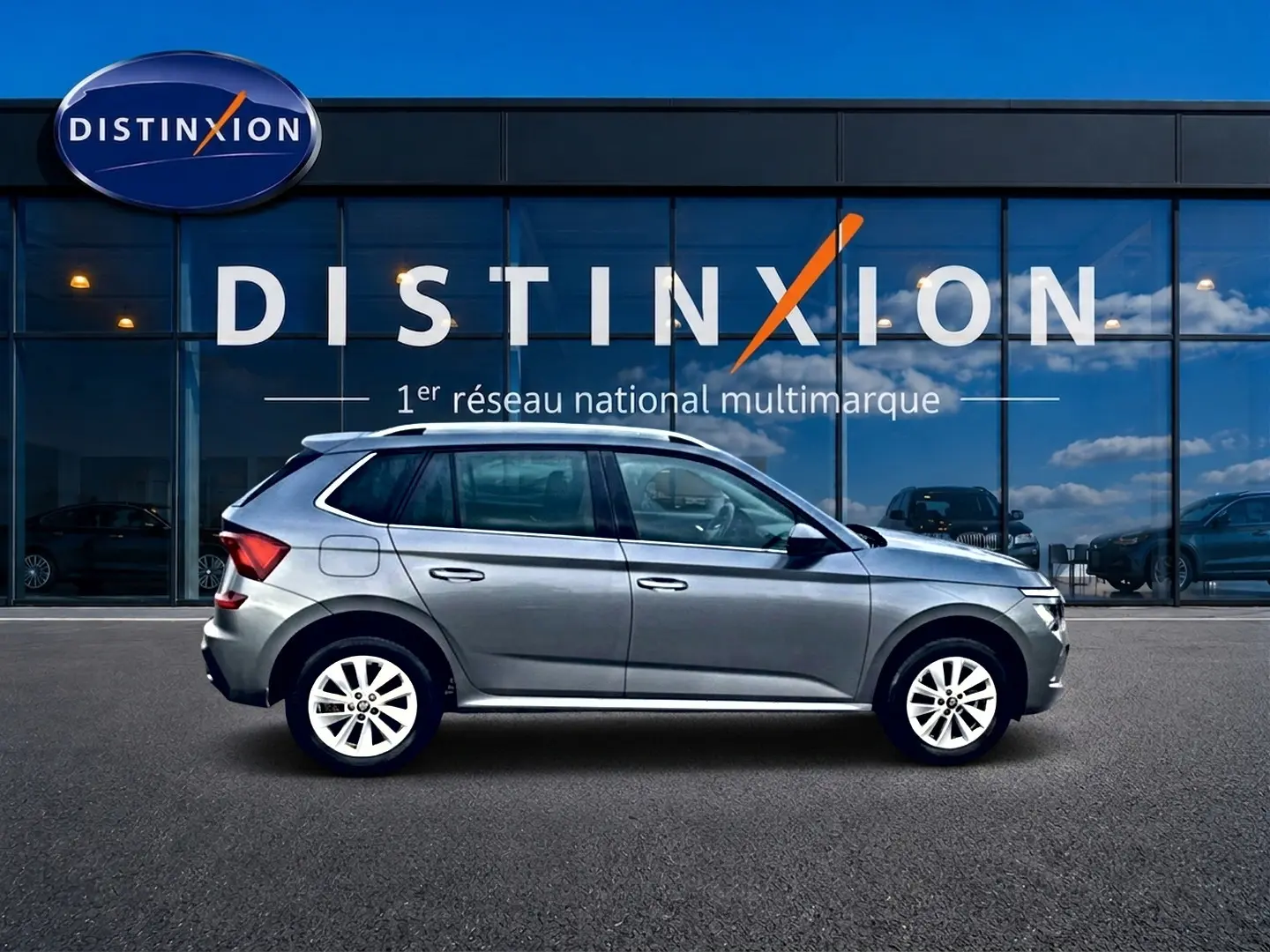 Vue de profil côté gauche d'un Skoda Kamiq gris 2025 garé devant une vitrine avec logo Distinxion.