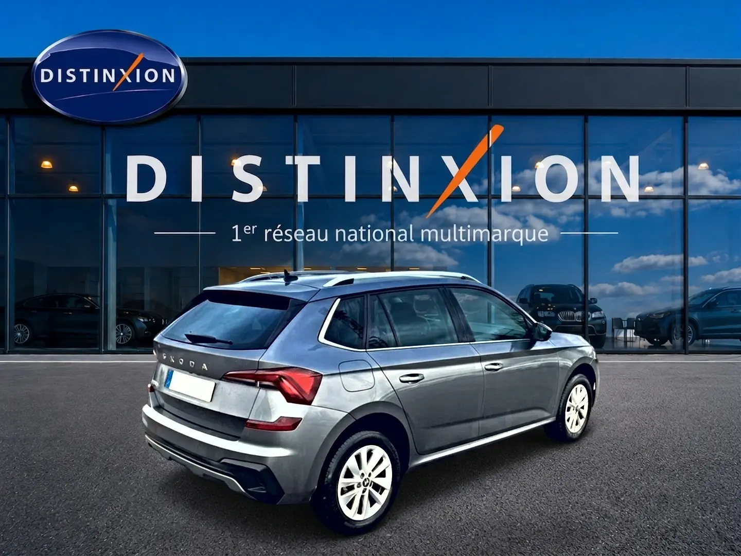 SKODA KAMIQ gris en 3/4 arrière droit, avec barres de toit et jantes alu blanches visibles devant un showroom Distinxion.