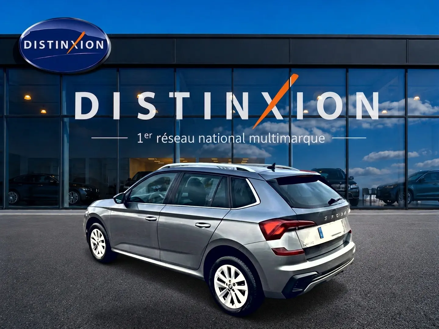 SKODA KAMIQ gris en 3/4 arrière droit, avec barres de toit et jantes alu blanches, devant une vitrine Distinxion.