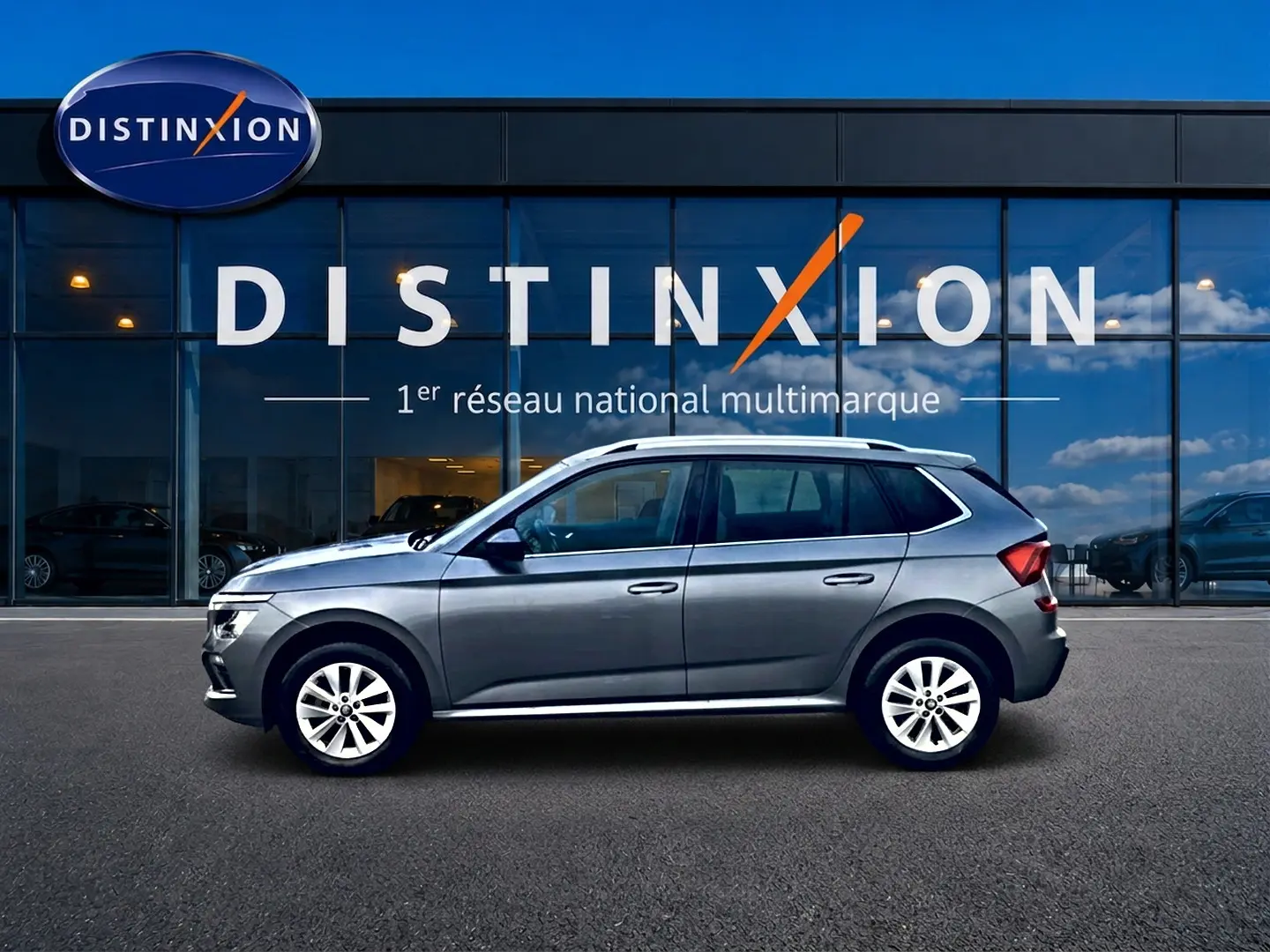 Vue latérale droite d'un Skoda Kamiq gris 2025 avec barres de toit et jantes alu devant une vitrine Distinxion.