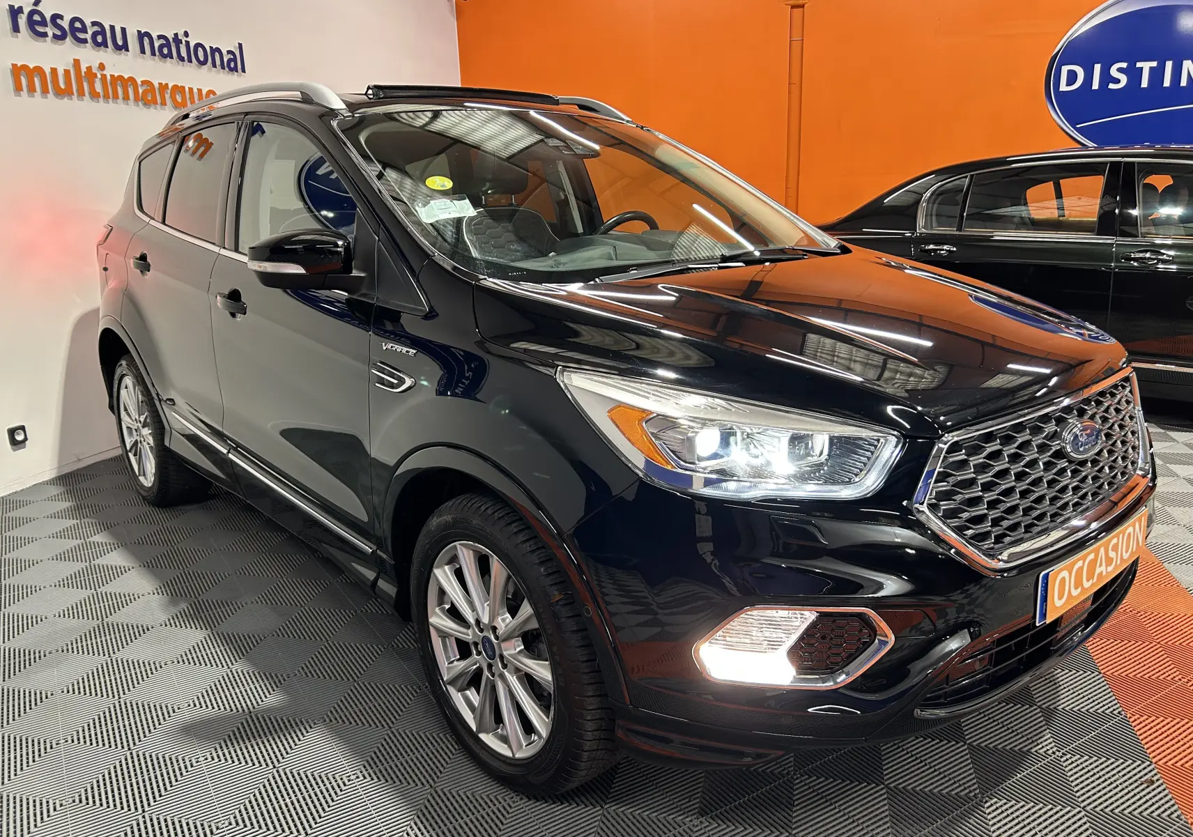 Ford Kuga noir en 3/4 avant droit, avec phares et feux antibrouillard allumés dans un showroom.