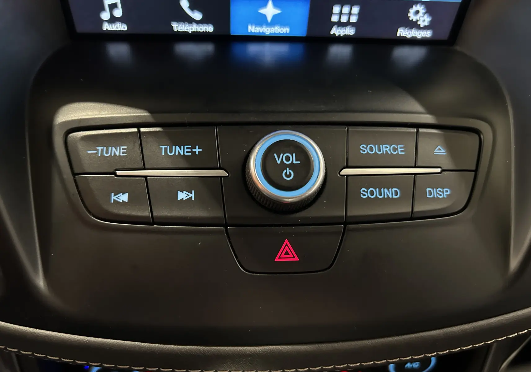 Gros plan sur la console centrale noire du Ford Kuga 2017, avec boutons de volume et commandes audio éclairés en bleu.