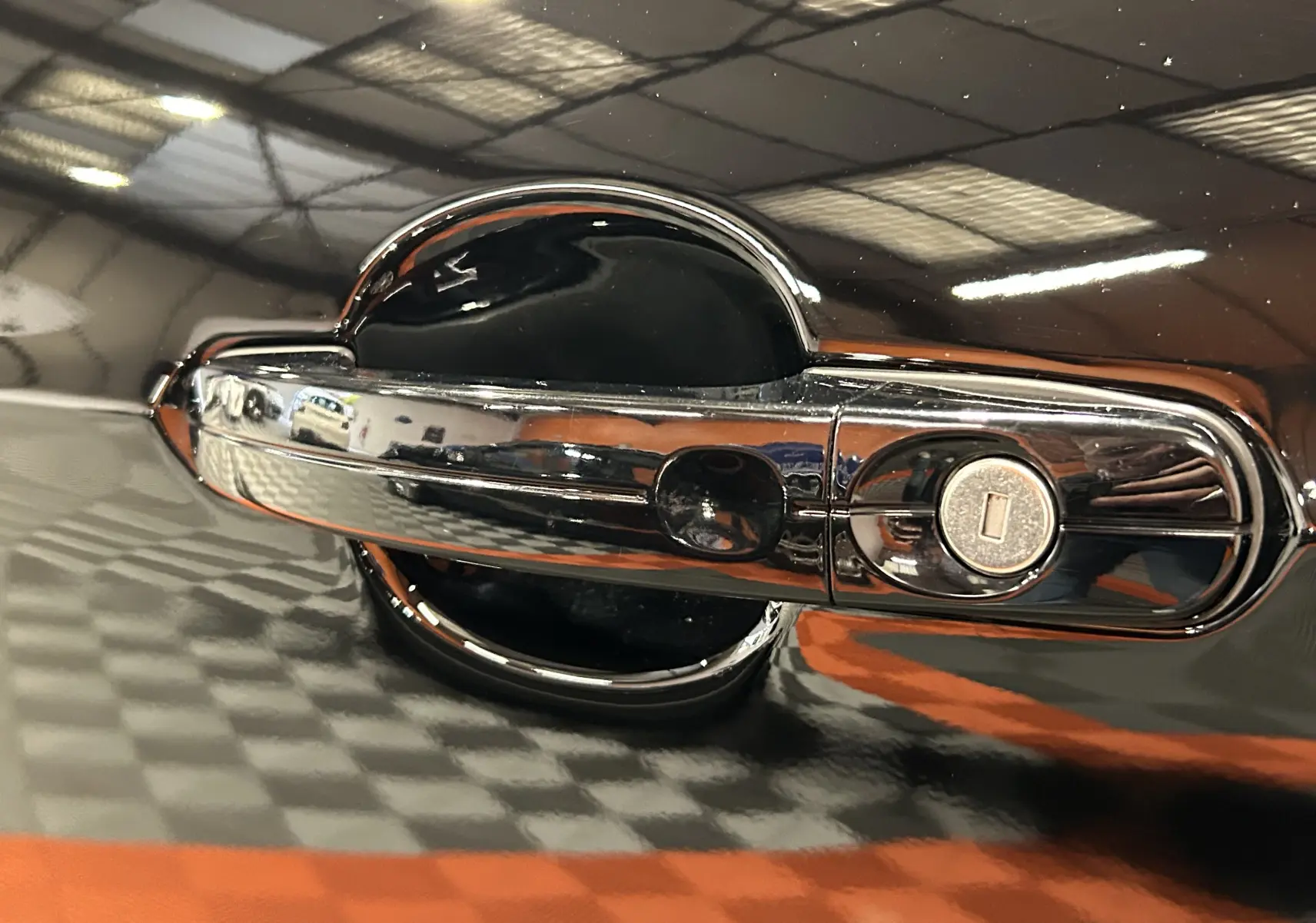Gros plan sur la poignée de porte chromée avec serrure d’un Ford Kuga noir, reflet du sol damier orange et gris visible.
