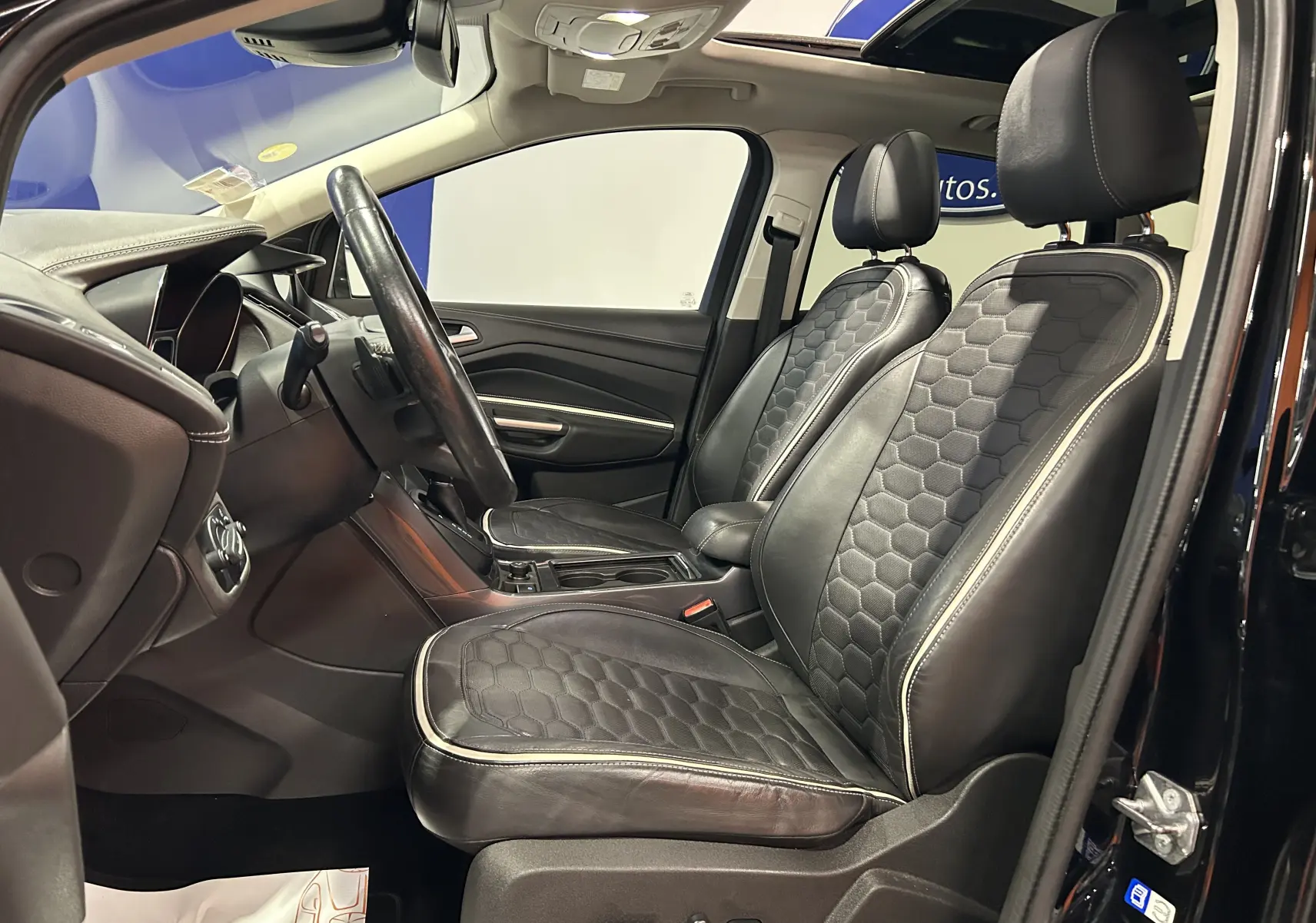 Vue intérieure côté conducteur du Ford Kuga noir 2017, sièges cuir noirs avec surpiqûres blanches et toit ouvrant panoramique.