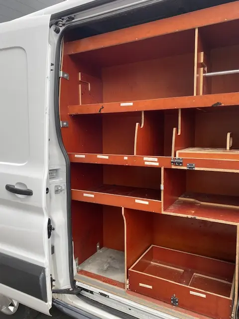 Vue partielle du fourgon Ford Transit blanc 2019, montrant l'intérieur aménagé avec étagères en bois côté droit ouvert.