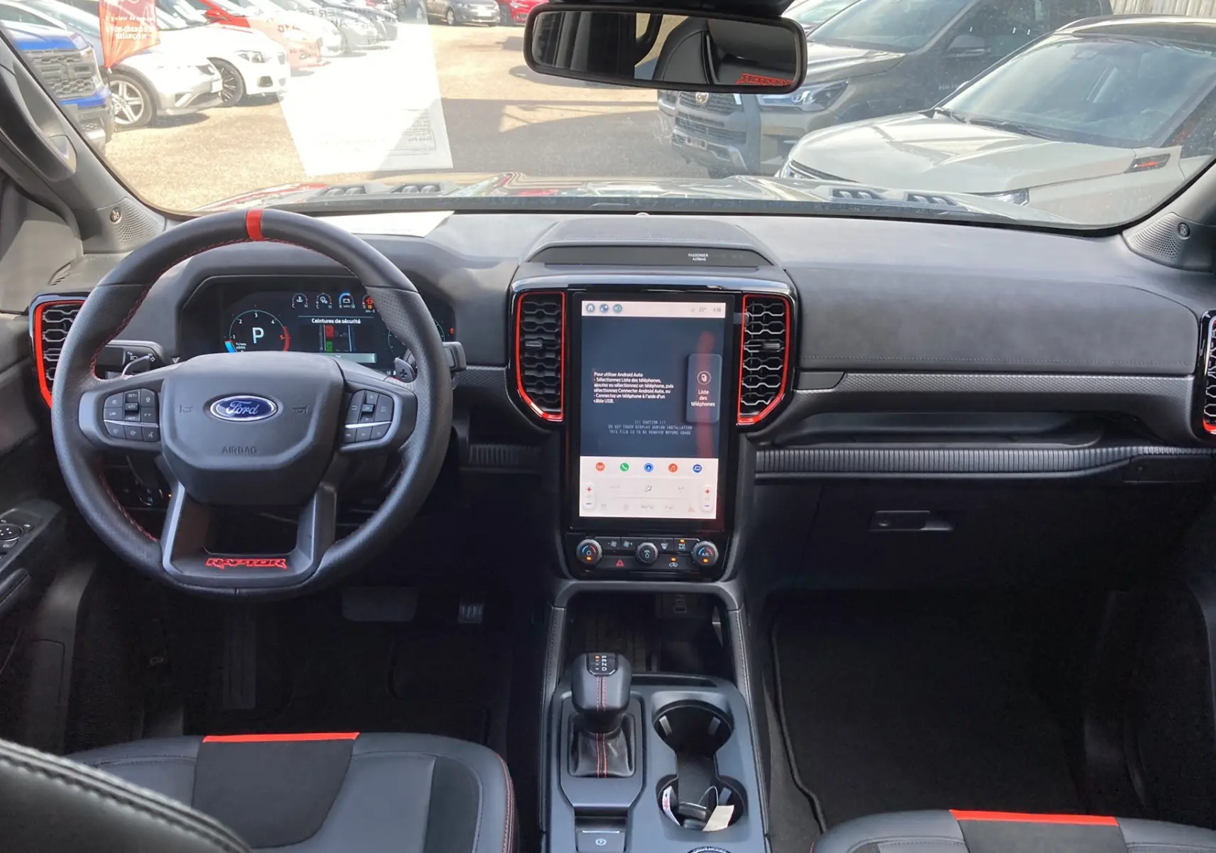 Intérieur avant du Ford Ranger Raptor 2026, vue du poste de conduite avec volant cuir noir et touches rouges, écran tactile central.