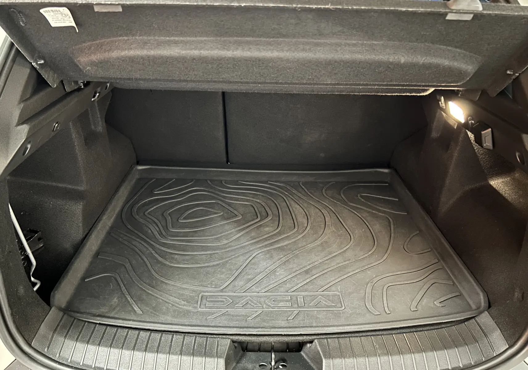 Coffre ouvert du Dacia Duster 2024 avec tapis de sol noir texturé et éclairage intérieur à droite.