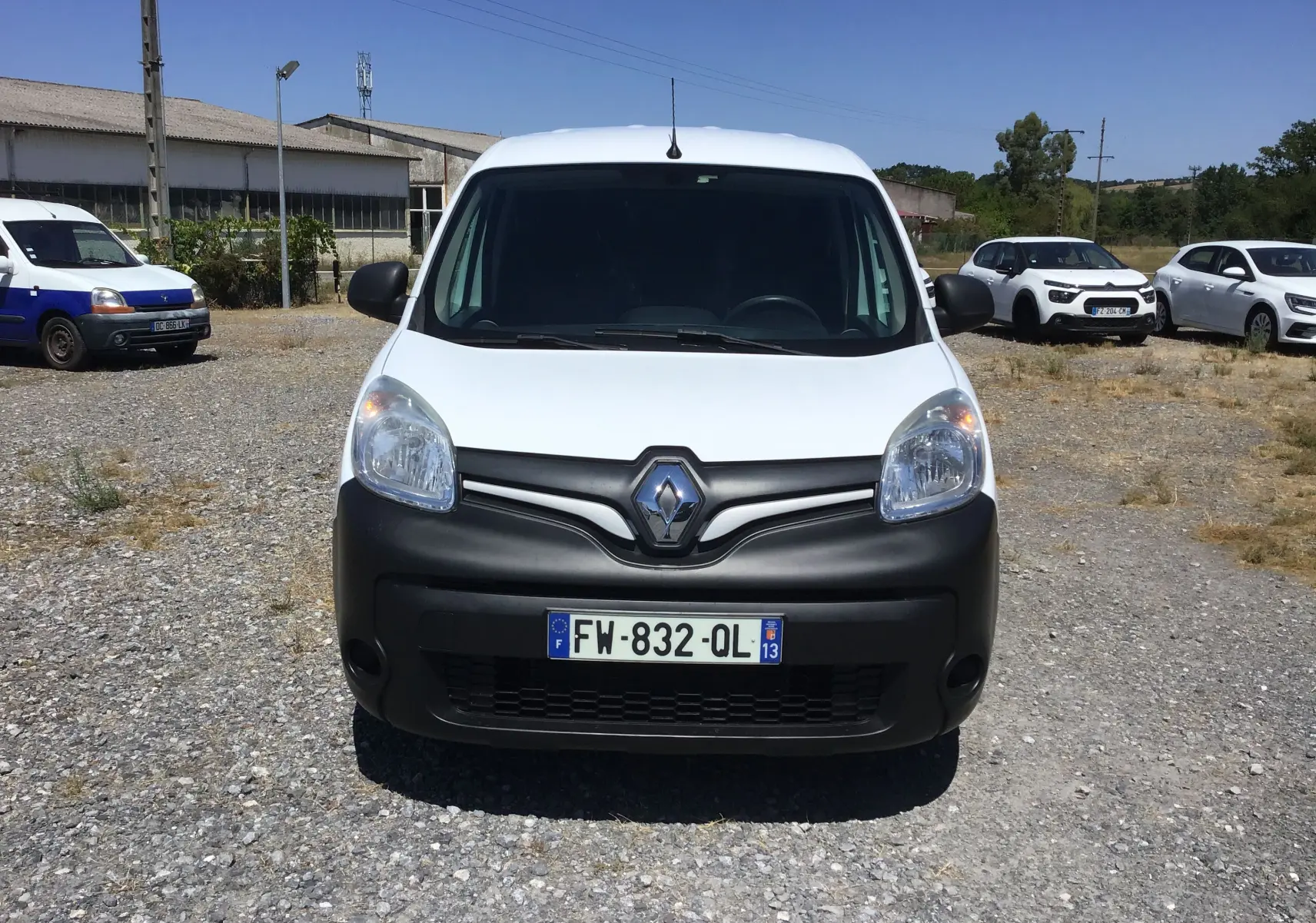 Vue de face d'un Renault Kangoo blanc avec pare-chocs noir mat, stationné sur un sol gravillonné en extérieur.