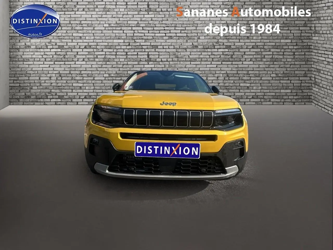 Vue frontale d'un Jeep Avenger jaune foncé avec calandre noire et logo Jeep visible sur fond de mur en briques.