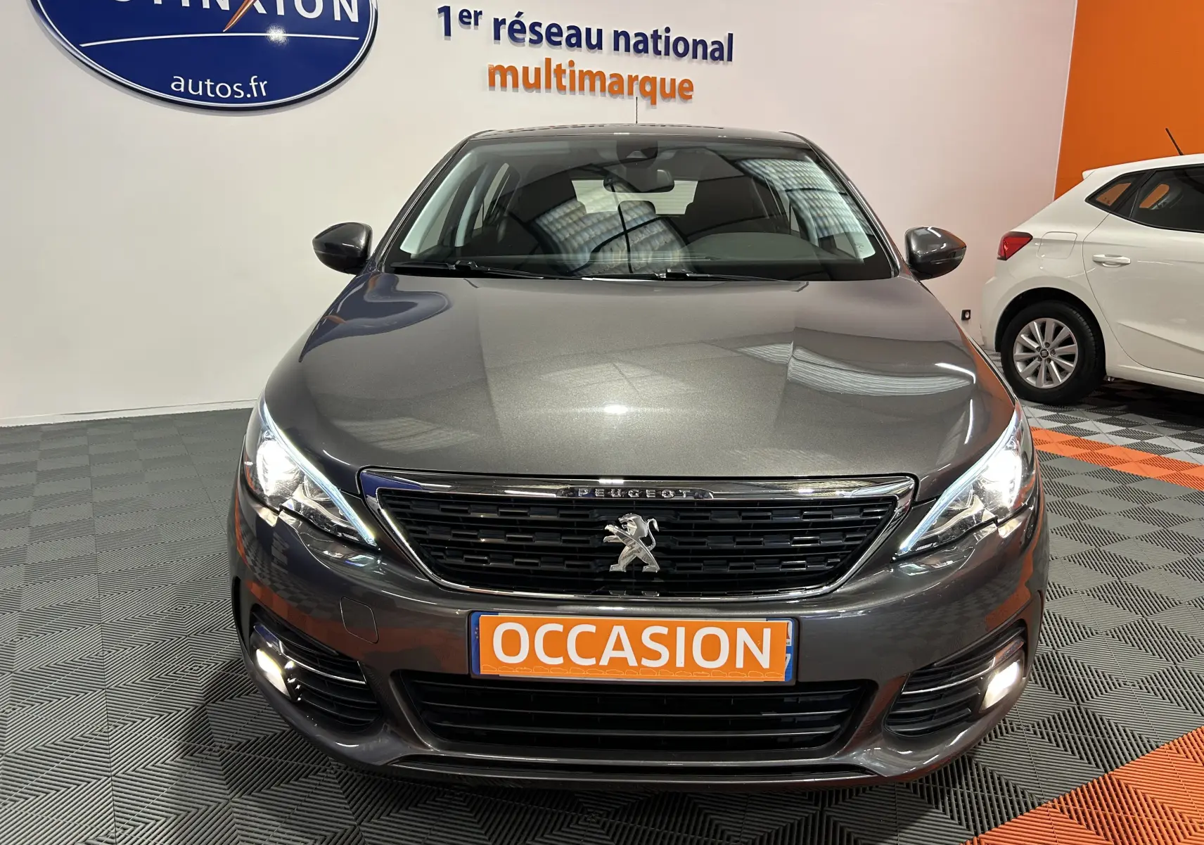 Peugeot 308 gris vue de face en intérieur, avec phares allumés et plaque orange "OCCASION" visible.